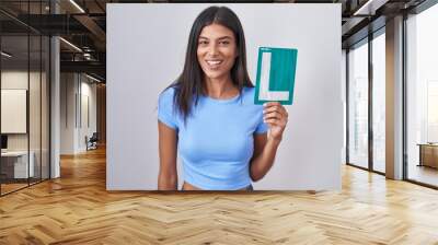 Brunette young woman holding l sign for new driver looking positive and happy standing and smiling with a confident smile showing teeth Wall mural