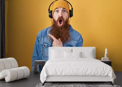 Caucasian man with long beard listening to music using headphones surprised pointing with finger to the side, open mouth amazed expression. Wall mural