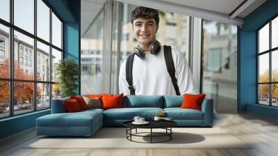 Cheerful young hispanic male student standing outdoors enjoying campus life, confidently wearing headphones and backpack, radiating positive energy and joy at the university. Wall mural