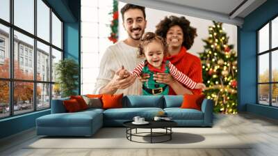 Couple and daughter looking to the camera standing by christmas tree at home Wall mural