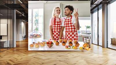 Couple of wife and husband cooking pastries at the kitchen showing and pointing up with fingers number two while smiling confident and happy. Wall mural