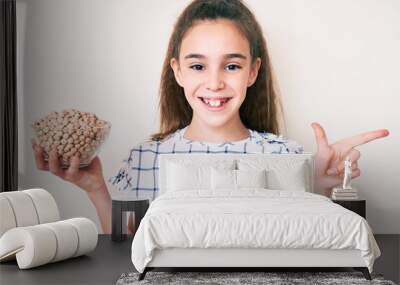 Cute hispanic child girl holding chickpeas bowl smiling happy pointing with hand and finger to the side Wall mural