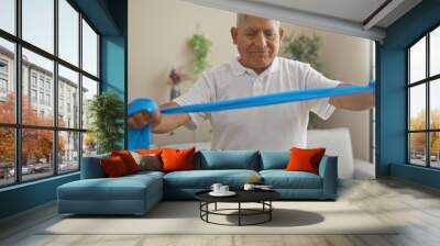 Elderly hispanic man exercising with a resistance band in a bright living room at home, focusing on fitness and wellness in a comfortable indoor setting. Wall mural