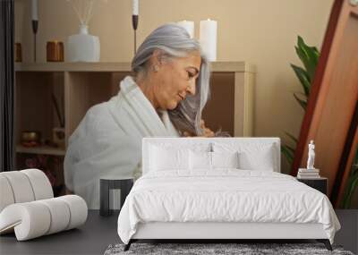 Elderly woman with grey hair in a spa room wearing a robe, gently touching her hair while standing in front of a mirror surrounded by candles and plants Wall mural