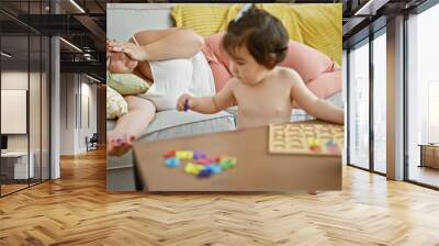 Exhausted mother and lovely daughter unwinding on the living room sofa, watching as baby joyously plays with educational math toys at home, cherishing time together Wall mural