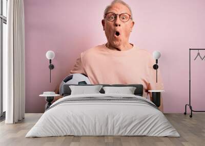 Grey haired senior football player man holding soccer ball over pink isolated background scared in shock with a surprise face, afraid and excited with fear expression Wall mural