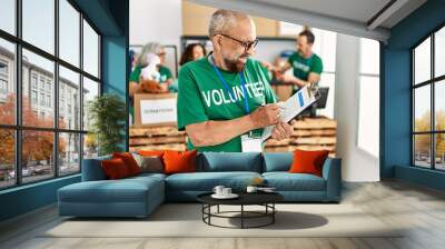 Group of middle age volunteers working at charity center. Man smiling happy writing on clipboard. Wall mural