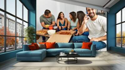 Group of young hispanic friends eating italian pizza sitting on the sofa. Man smiling happy and talking on the smartphone at home. Wall mural