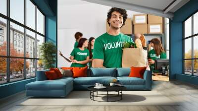 Group of young volunteers working at charity center. Man smiling happy and holding paper bag with food to donate. Wall mural