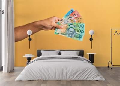 Hand of hispanic man holding australian dollars banknotes over isolated yellow background. Wall mural