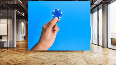 hand of hispanic man holding casino chip over isolated blue background. Wall mural