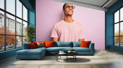 Handsome african american man wearing casual t-shirt and glasses over pink background looking away to side with smile on face, natural expression. Laughing confident. Wall mural