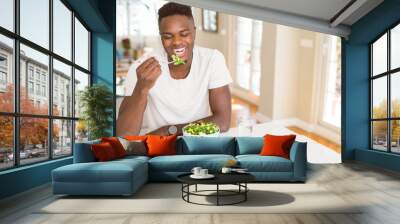 Handsome african young man eating a healthy vegetable salad using a fork to eat lettuce, happy and smiling sitting on the table Wall mural