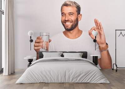 Handsome blond man with beard drinking glass of water to refreshment over white background doing ok sign with fingers, smiling friendly gesturing excellent symbol Wall mural