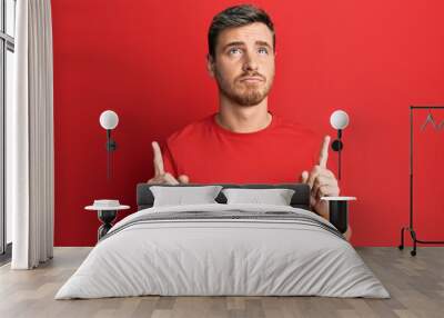 Handsome caucasian man wearing casual red tshirt pointing up looking sad and upset, indicating direction with fingers, unhappy and depressed. Wall mural