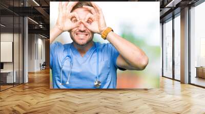 Handsome doctor man wearing medical uniform over isolated background doing ok gesture like binoculars sticking tongue out, eyes looking through fingers. Crazy expression. Wall mural