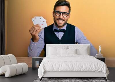 Handsome hispanic croupier man sitting on the table with poker chips and cards looking positive and happy standing and smiling with a confident smile showing teeth Wall mural