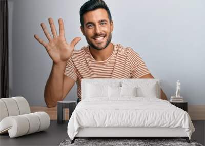 Handsome hispanic man wearing casual clothes sitting on the table showing and pointing up with fingers number five while smiling confident and happy. Wall mural