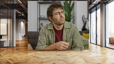 Handsome hispanic man with beard and glasses using wireless earphones at home, reflecting a casual lifestyle in a cozy interior. Wall mural