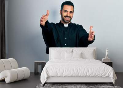 Handsome hispanic man with beard wearing catholic priest robe looking at the camera smiling with open arms for hug. cheerful expression embracing happiness. Wall mural