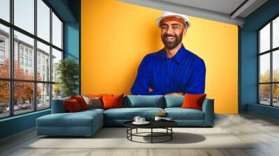 Handsome indian worker man wearing uniform and helmet over isolated yellow background happy face smiling with crossed arms looking at the camera. Positive person. Wall mural