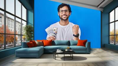 Handsome latin man holding dollars banknotes screaming proud, celebrating victory and success very excited with raised arms Wall mural