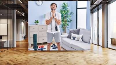 Handsome latin man wearing sportswear at home praying with hands together asking for forgiveness smiling confident. Wall mural