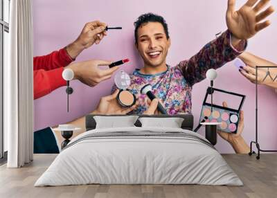 Handsome man wearing make up with make up cosmetics around looking at the camera smiling with open arms for hug. cheerful expression embracing happiness. Wall mural