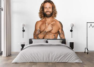 Handsome man with beard and long hair standing shirtless showing tattoos cheerful with a smile on face pointing with hand and finger up to the side with happy and natural expression Wall mural