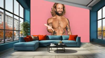 Handsome man with beard and long hair standing shirtless showing tattoos smiling cheerful offering palm hand giving assistance and acceptance. Wall mural