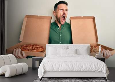 Handsome man with beard holding two italian pizzas angry and mad screaming frustrated and furious, shouting with anger. rage and aggressive concept. Wall mural