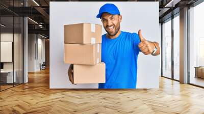 Handsome man with beard wearing courier uniform holding delivery packages approving doing positive gesture with hand, thumbs up smiling and happy for success. winner gesture. Wall mural