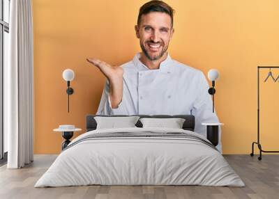 Handsome man with beard wearing professional cook uniform smiling cheerful presenting and pointing with palm of hand looking at the camera. Wall mural