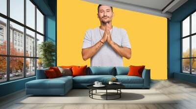 Handsome man with tattoos wearing 90s style praying with hands together asking for forgiveness smiling confident. Wall mural