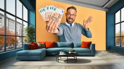 Handsome middle age man holding bunch of euros celebrating achievement with happy smile and winner expression with raised hand Wall mural