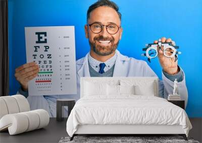 Handsome middle age man holding optometry glasses and medical exam smiling with a happy and cool smile on face. showing teeth. Wall mural