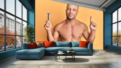 Handsome muscle man holding razor smiling with an idea or question pointing finger with happy face, number one Wall mural