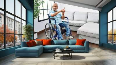 Handsome senior man sitting on wheelchair at the living room smiling and looking at the camera pointing with two hands and fingers to the side. Wall mural