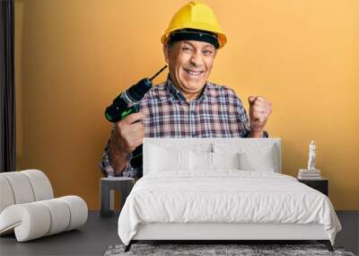 Handsome senior man with grey hair holding electric screwdriver wearing hardhat screaming proud, celebrating victory and success very excited with raised arm Wall mural