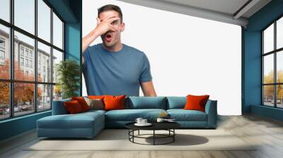 Handsome young casual man peeking in shock covering face and eyes with hand, looking through fingers with embarrassed expression. Wall mural