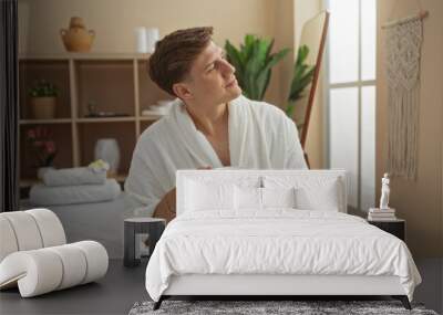 Handsome young man enjoying a relaxing moment at a wellness spa, dressed in a white robe and holding a blue cup of tea, surrounded by a cozy interior with plants and soft lighting Wall mural