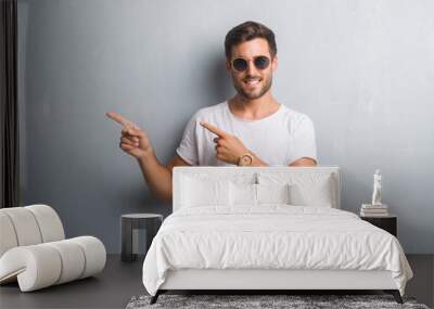 Handsome young man over grey grunge wall wearing sunglasses smiling and looking at the camera pointing with two hands and fingers to the side. Wall mural