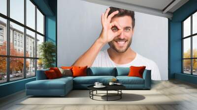 Handsome young man over grey grunge wall with happy face smiling doing ok sign with hand on eye looking through fingers Wall mural