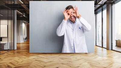 Handsome young professional man over grey grunge wall wearing white coat doing ok gesture like binoculars sticking tongue out, eyes looking through fingers. Crazy expression. Wall mural