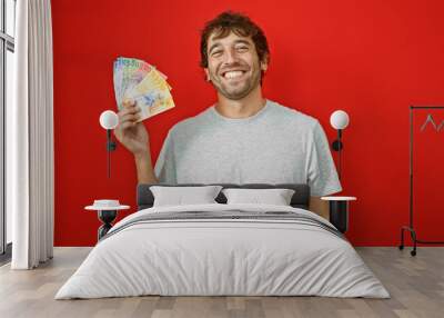 Happy young man with blond beard proudly holding up swiss franc banknotes, financial triumph writ large on his face, depicted in an isolated, vivacious red background portrait! Wall mural