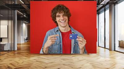 Happy young man with curly hair pointing at credit card against vibrant red background Wall mural