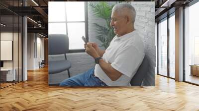 Hispanic, grey-haired man seated indoors in a waiting room, using a phone with a smile on his face, featuring a modern, white brick interior and green plants. Wall mural