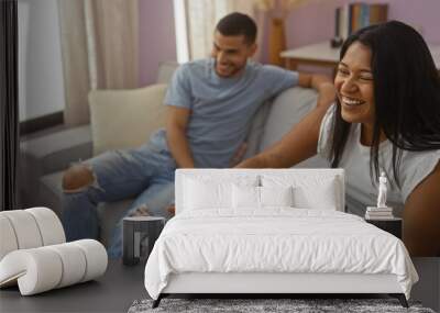 Hispanic couple laughing together in a cozy living room, sitting on a sofa and enjoying each other's company indoors. Wall mural