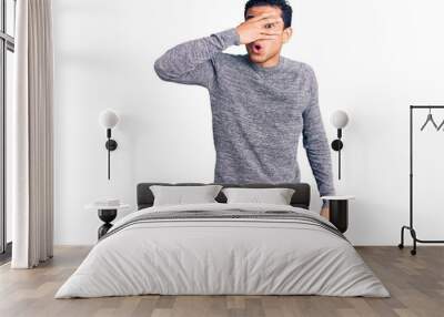 Hispanic handsome young man wearing casual sweater peeking in shock covering face and eyes with hand, looking through fingers with embarrassed expression. Wall mural