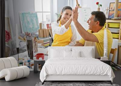 Hispanic man and woman artists high five in joy at art studio while drawing on notebook; vibrant celebration amidst paintbrushes, easels, and canvas around Wall mural